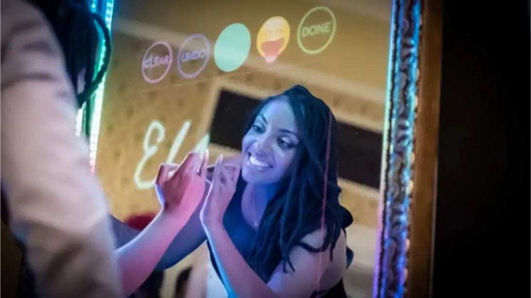 Interactive touchscreen mirror photo booth with a smiling guest engaging with the display, showcasing colorful interface options for capturing memorable moments at events.