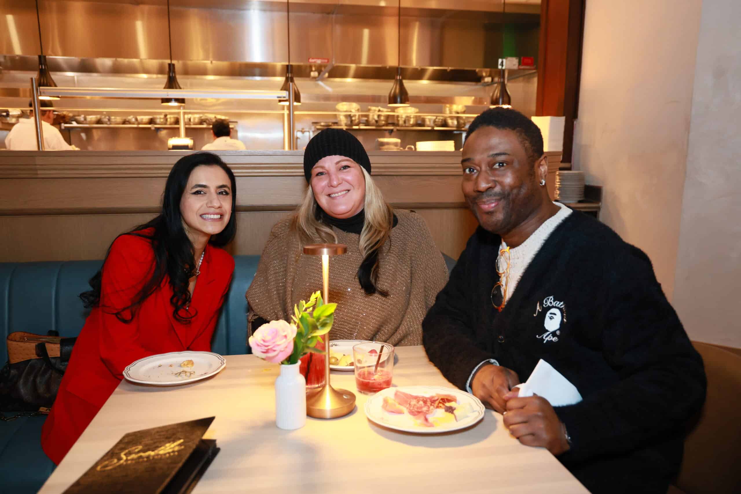 Three guests enjoying cocktails during a cocktail hour, seated at a table with plates of food and a floral centerpiece, in a lively restaurant setting.