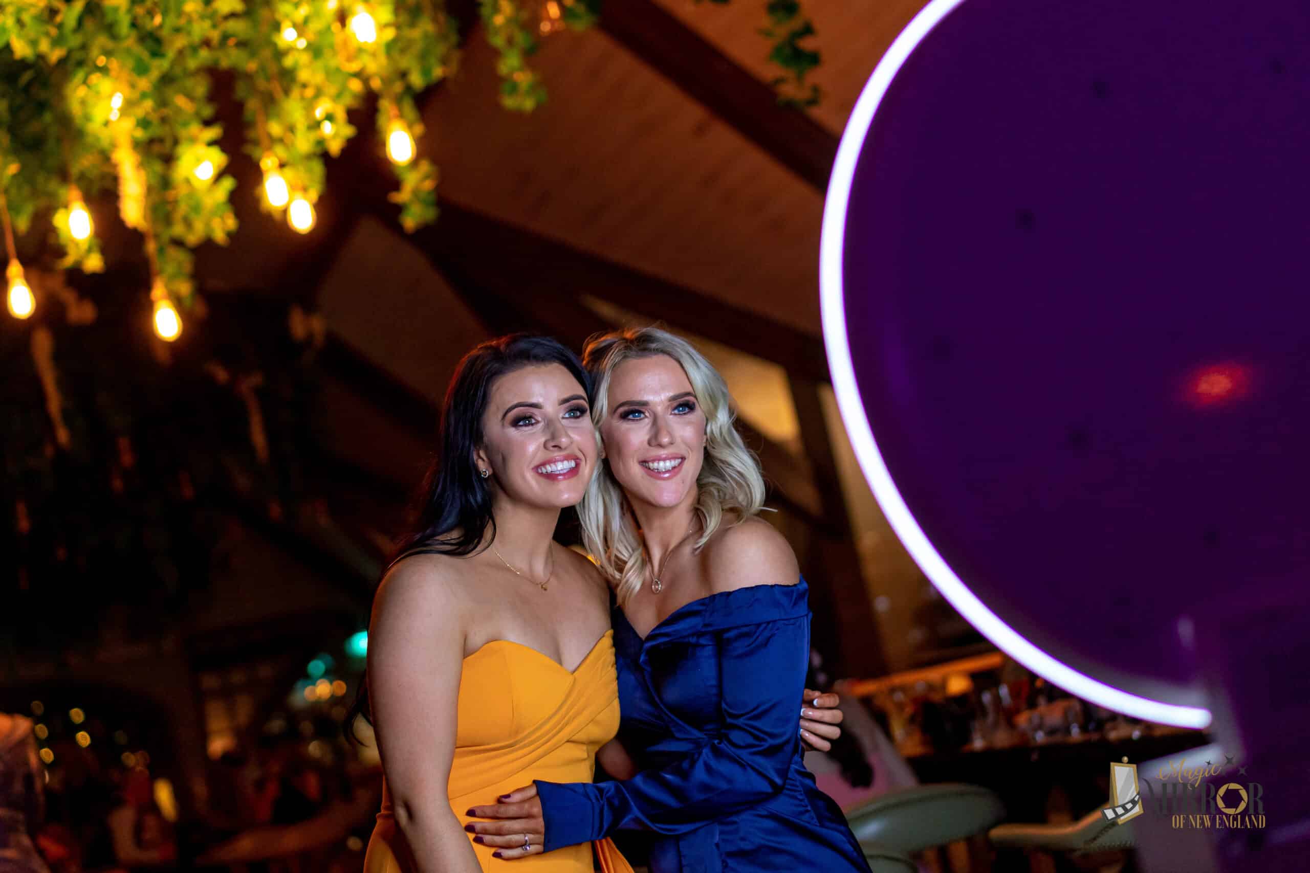 Two women smiling and posing together in a vibrant event setting, illuminated by warm lights, with a photo booth featuring a circular light in the foreground, capturing a joyful moment at a private celebration.
