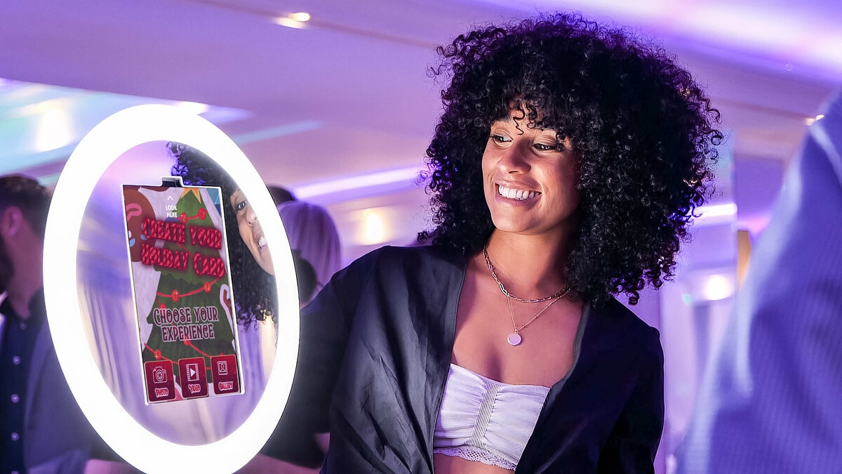 Interactive photo booth experience featuring a woman holding a mirror with a holiday card creation prompt, set in a festive event atmosphere.