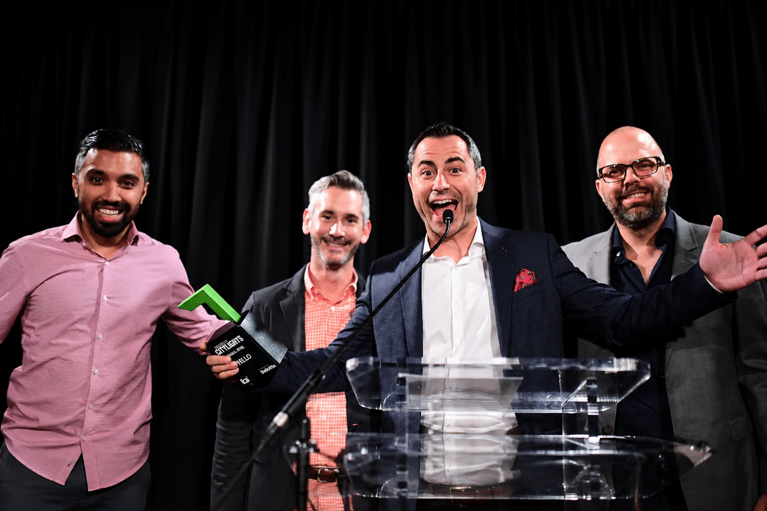 Four men celebrating on stage at a corporate awards ceremony, one holding a green trophy at a podium with a microphone, capturing an energetic recognition moment.