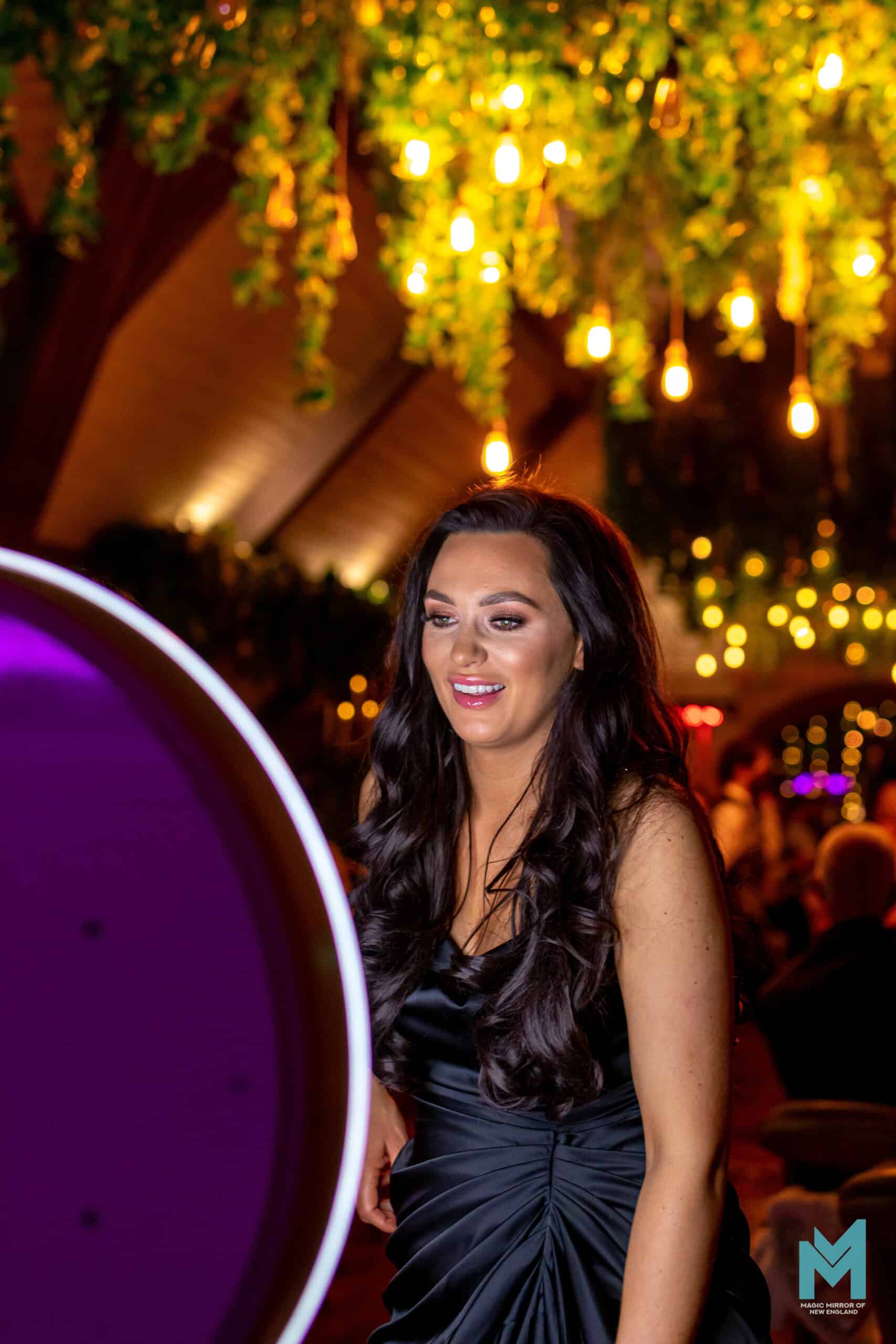 Guest in a dark dress smiling beside the Selfie Station Luxe photo booth under hanging greenery and string lights