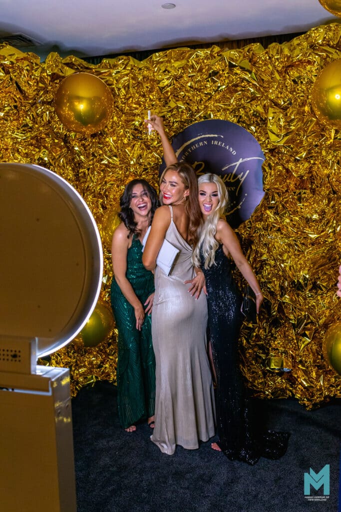 Three guests in evening wear posing in front of a gold backdrop next to the Selfie Station Luxe photo booth