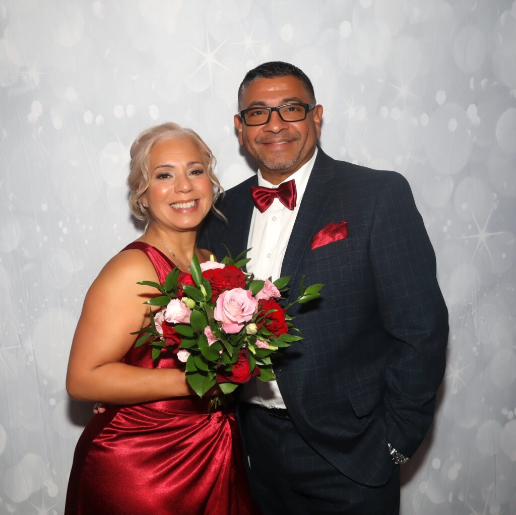 Couple posing with a red and pink bouquet in front of a silver sparkle backdrop, photographed on the Selfie Station Luxe