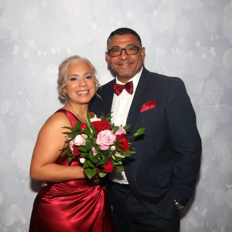 Couple posing with a red and pink bouquet in front of a silver sparkle backdrop, photographed on the Selfie Station Luxe
