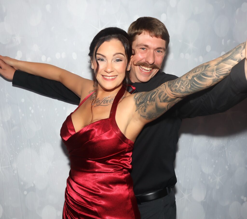 Couple posing against a silver bokeh backdrop, the woman in a red satin dress with her arms stretched out