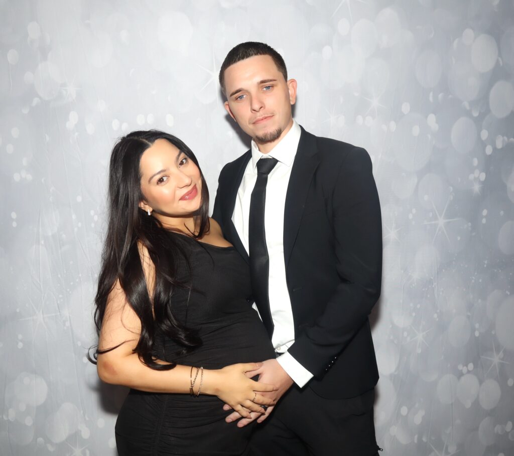 Couple in black formal outfits posing against a silver bokeh backdrop