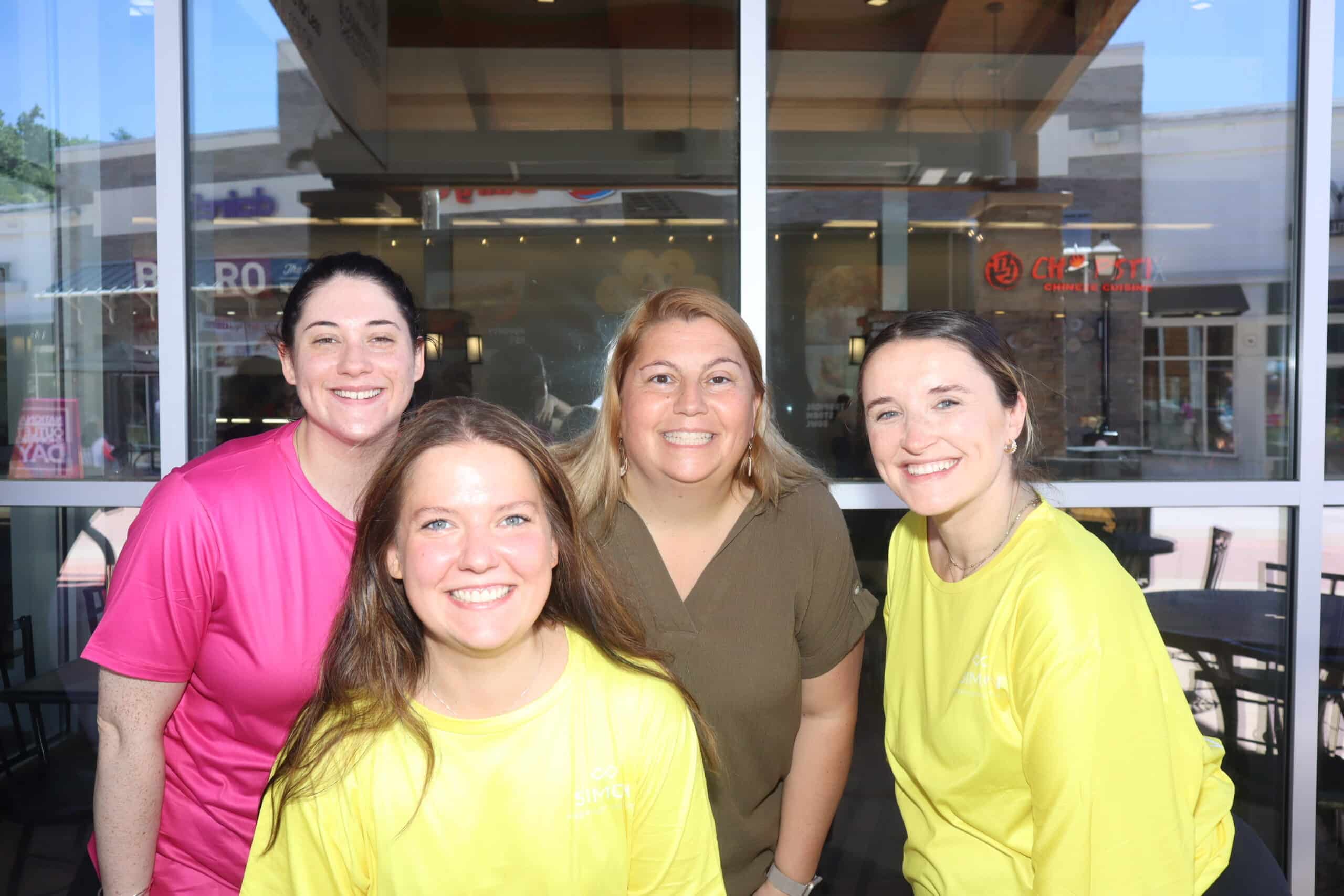 Four smiling women posing together outside a storefront during the day