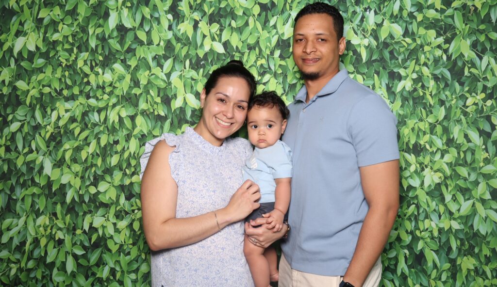 Family of three posing together at a photo booth with a greenery backdrop