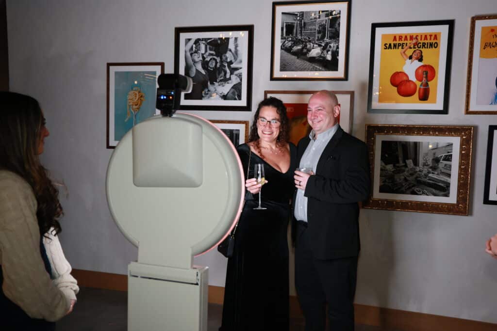 Two guests posing beside a white Selfie Station Luxe open-air photo booth with a professional camera, in front of a wall of framed art