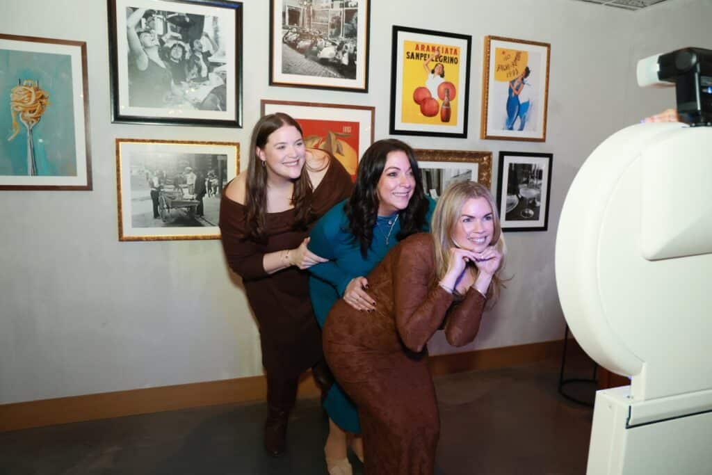 Three friends posing playfully beside the Selfie Station Luxe photo booth in front of a gallery wall of framed art
