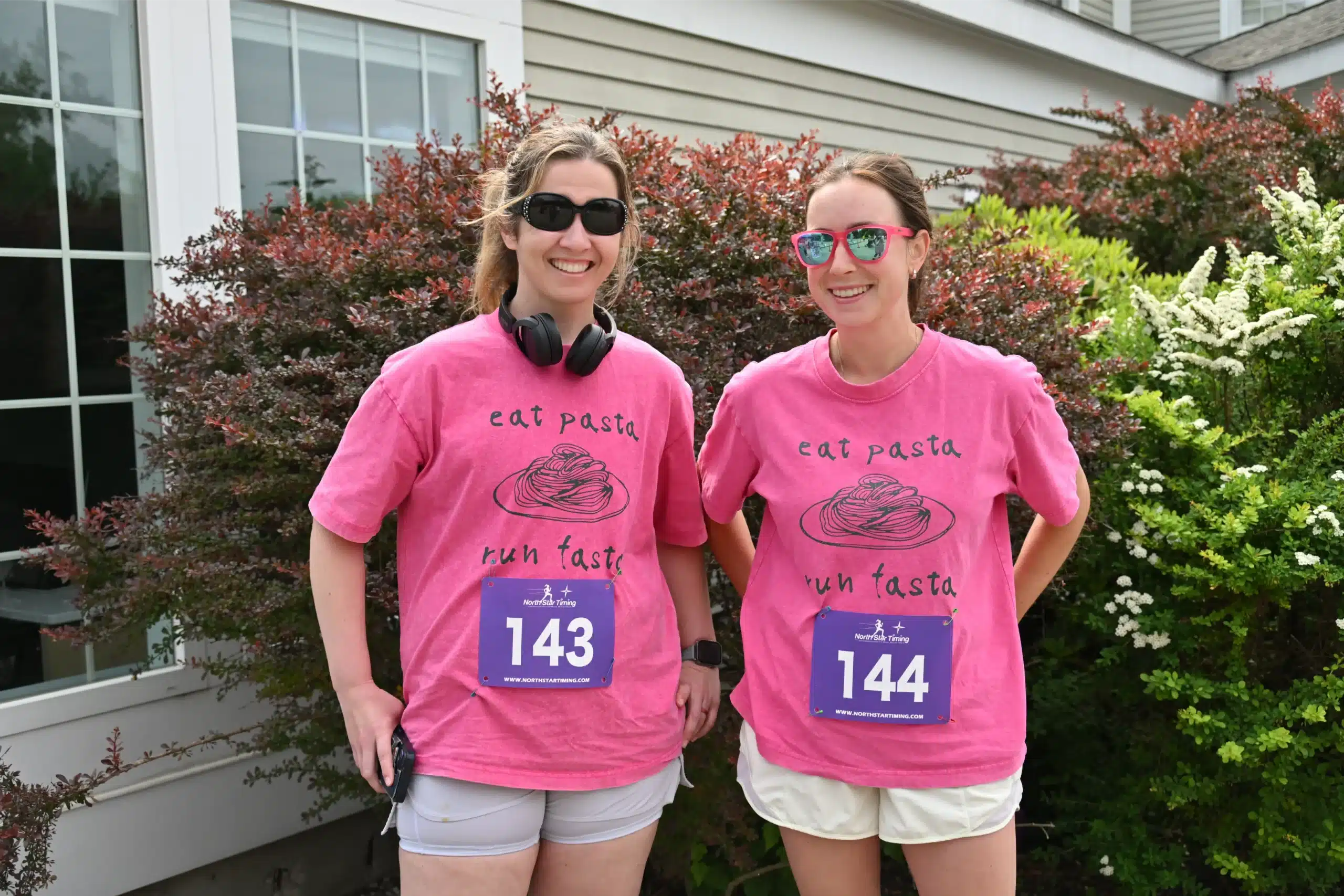 Two women in matching pink race t-shirts and bib numbers posing outdoors before a run