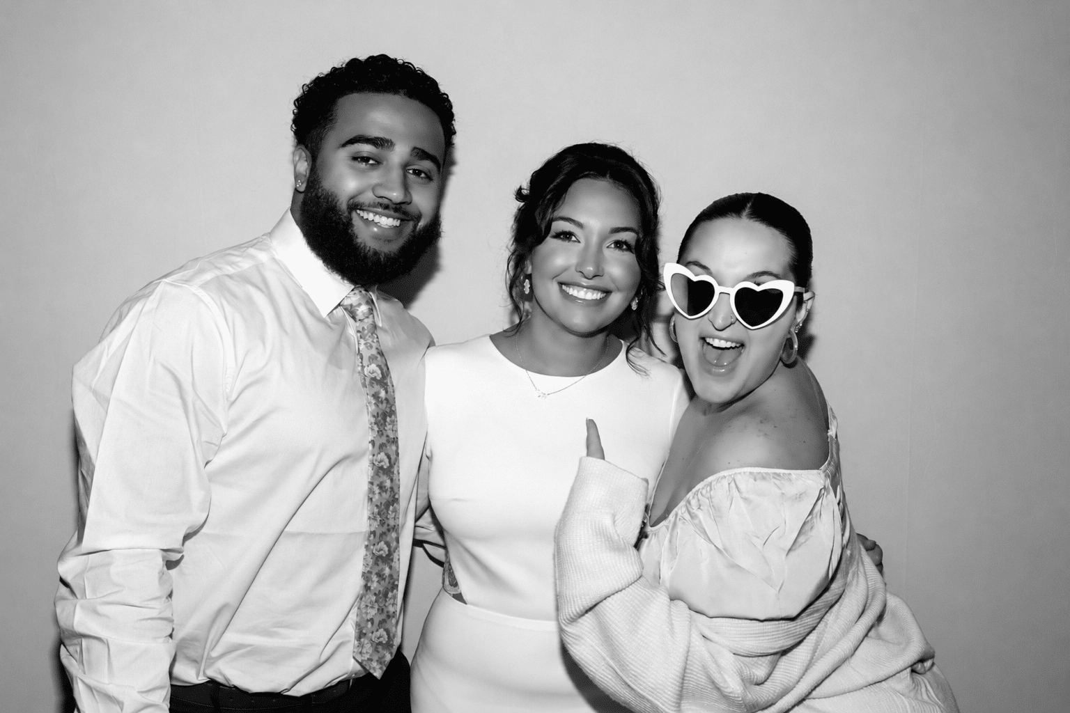 Three wedding guests posing together in a black and white photo booth portrait, with one guest wearing heart-shaped sunglasses.