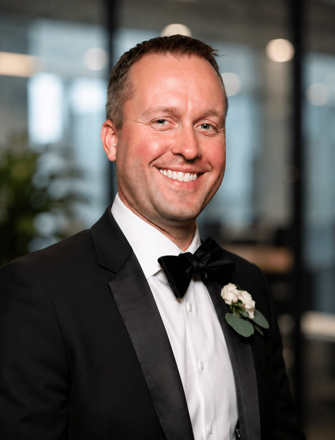 Man in a tuxedo with a bow tie and boutonnière, smiling in a modern event setting, representing professional AI headshot photography for corporate events and gatherings.