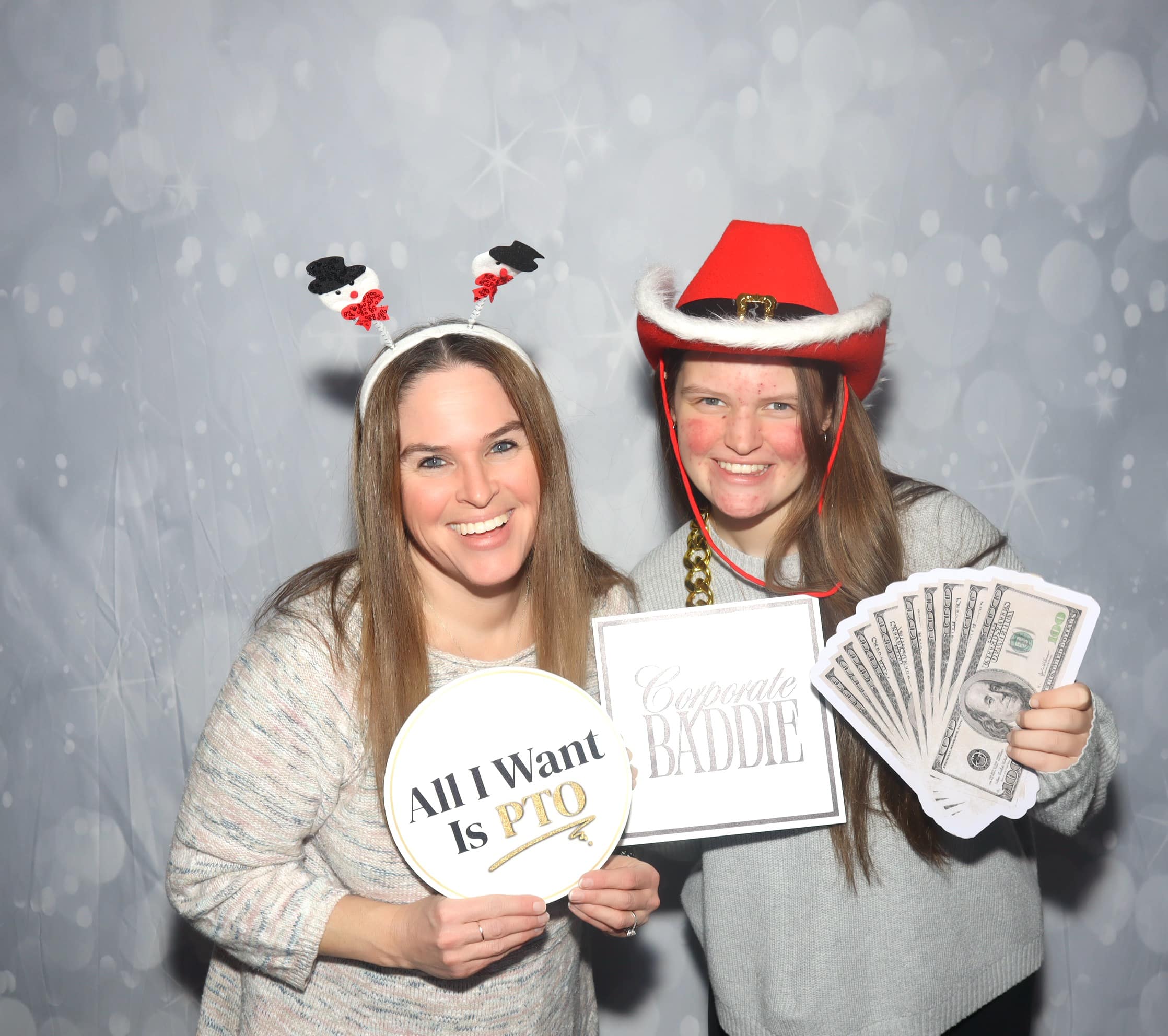 Two women posing at a holiday photo booth with festive props and humorous prop signs