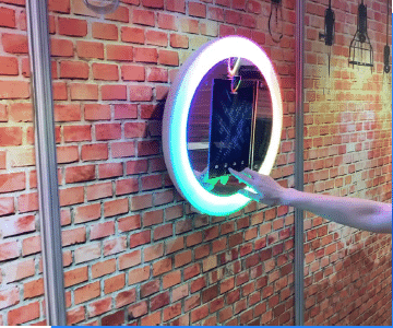 Wall-mounted circular photo booth mirror with illuminated LED ring, installed against an exposed brick wall as a permanent venue installation