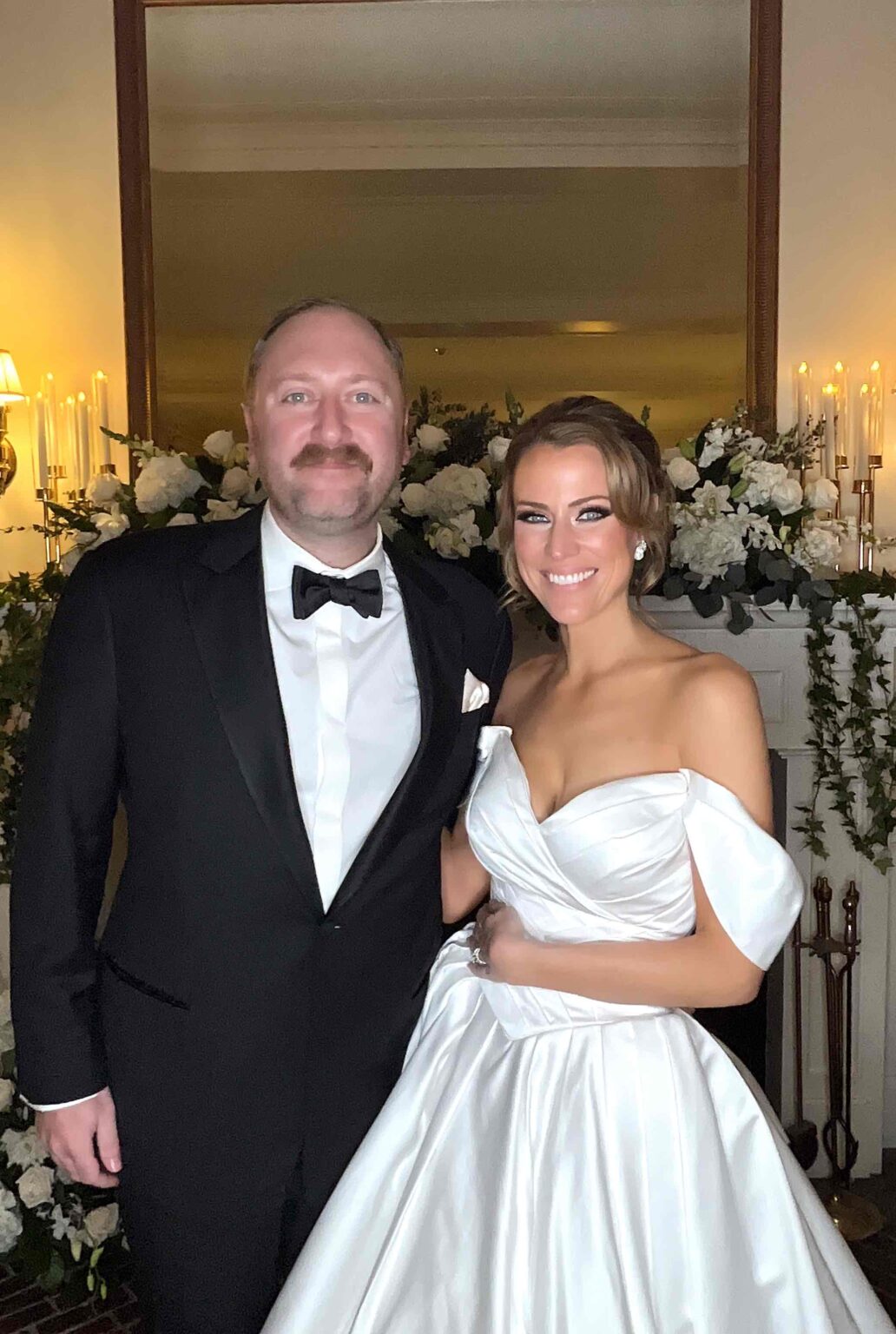 A bride in a white wedding gown and a groom in a tuxedo posing in front of a flower wall at their wedding