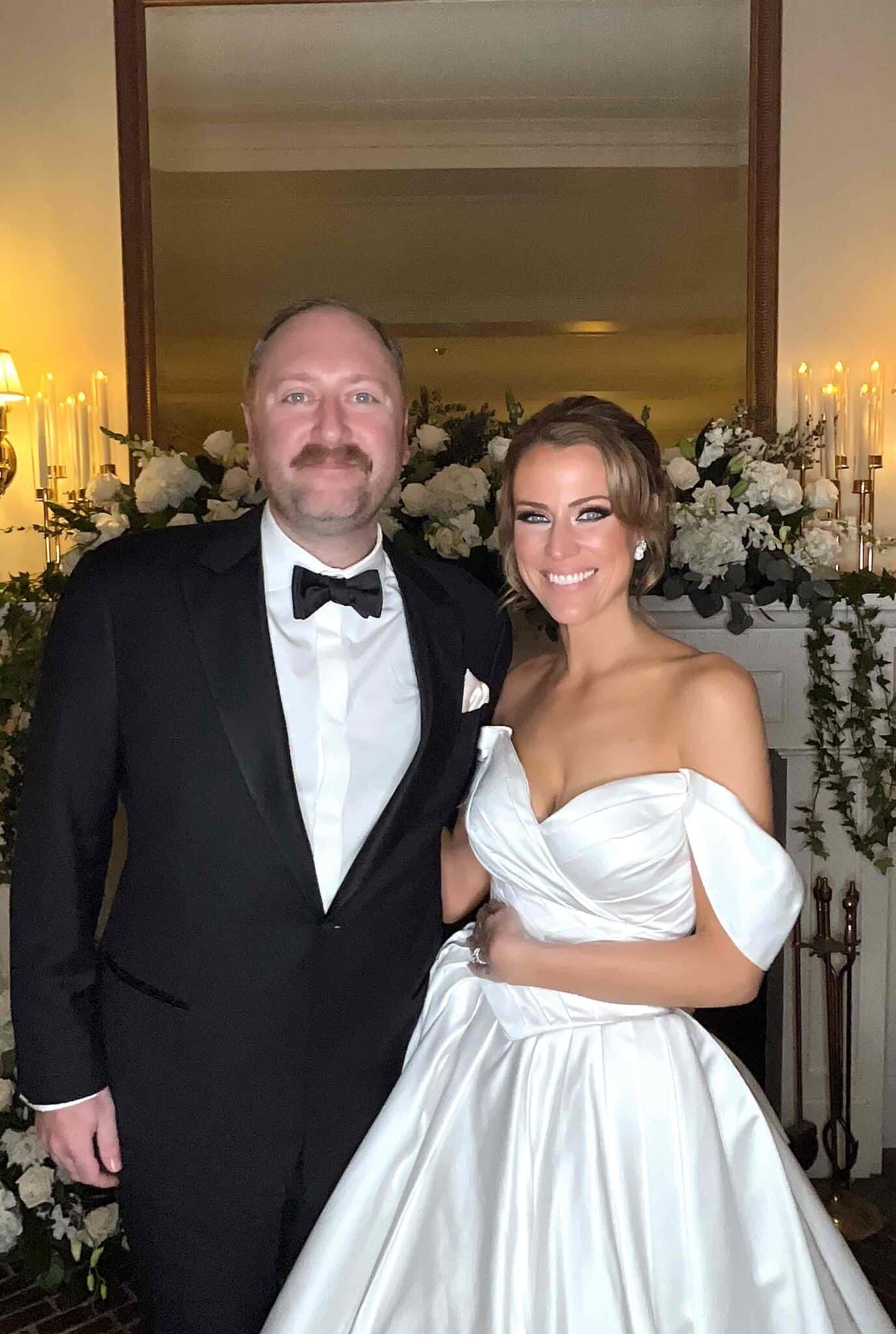 A bride in a white wedding gown and a groom in a tuxedo posing in front of a flower wall at their wedding