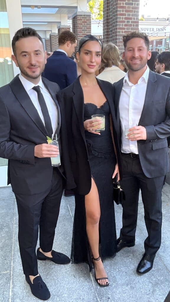 Three guests in formal wear holding cocktails on the terrace at a wedding cocktail hour with a roaming photo booth in NH