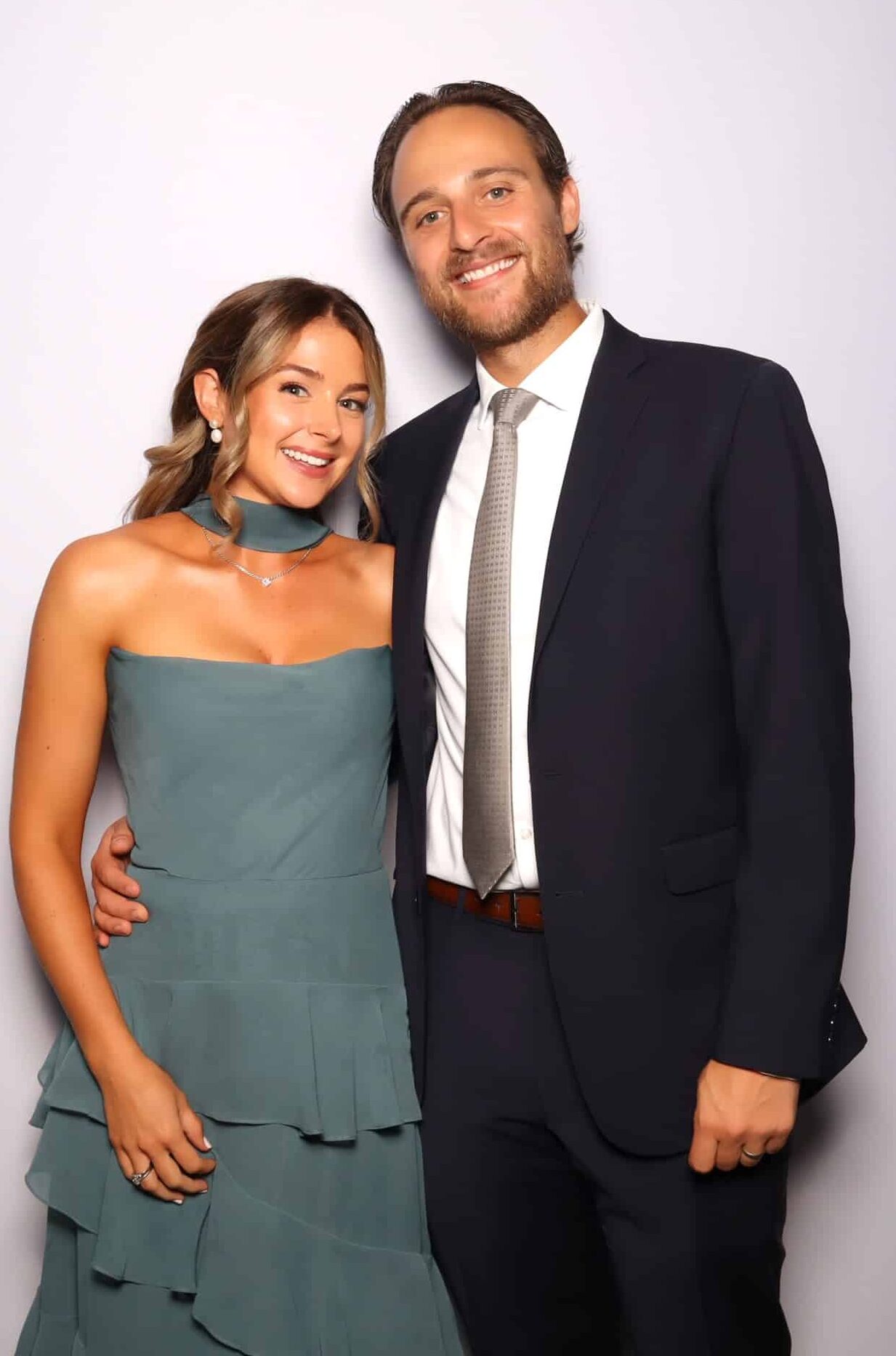 Couple posing together in front of a light backdrop, the woman in a teal dress and the man in a navy suit