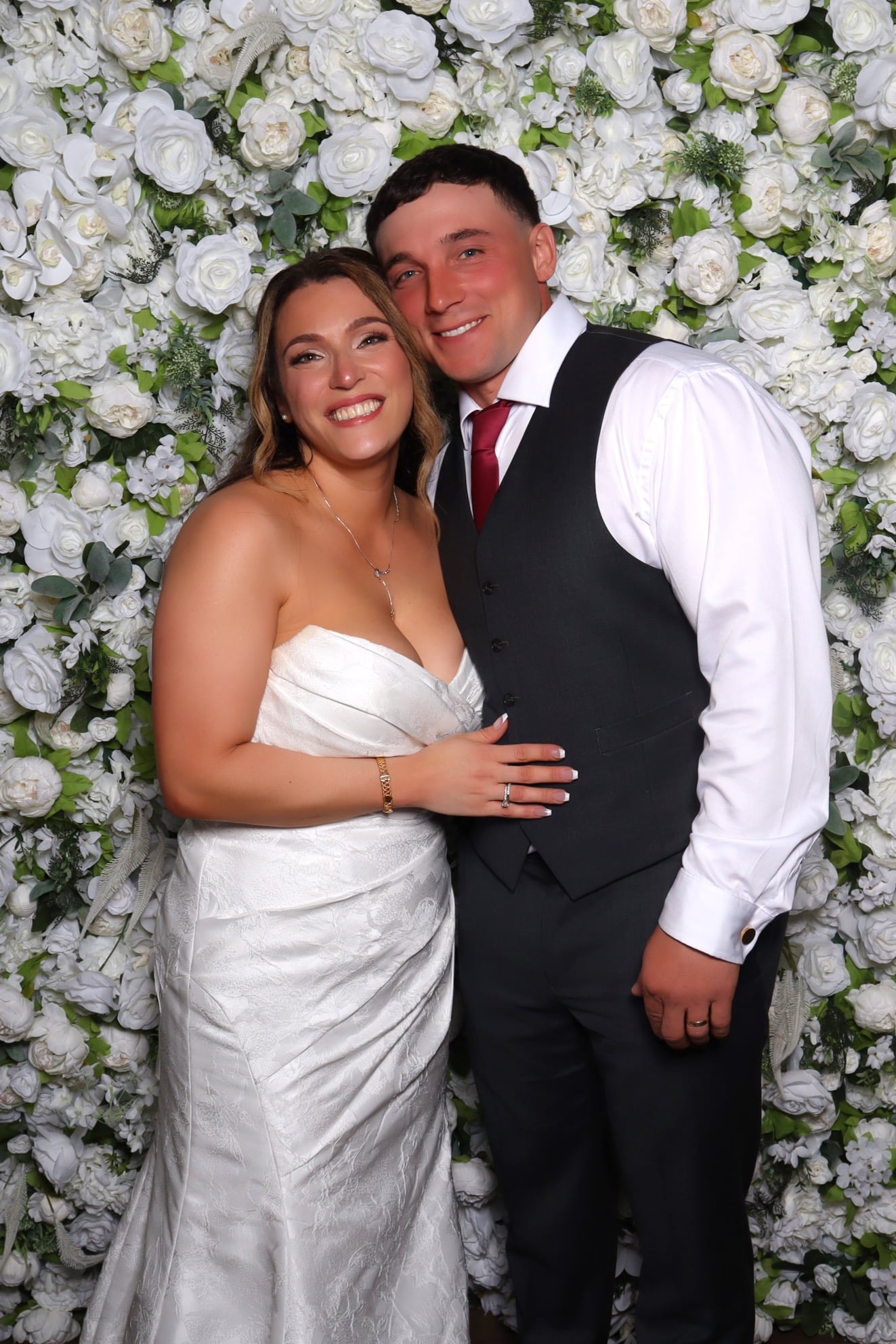Couple in a white dress and dark vest posing in front of a white flower wall