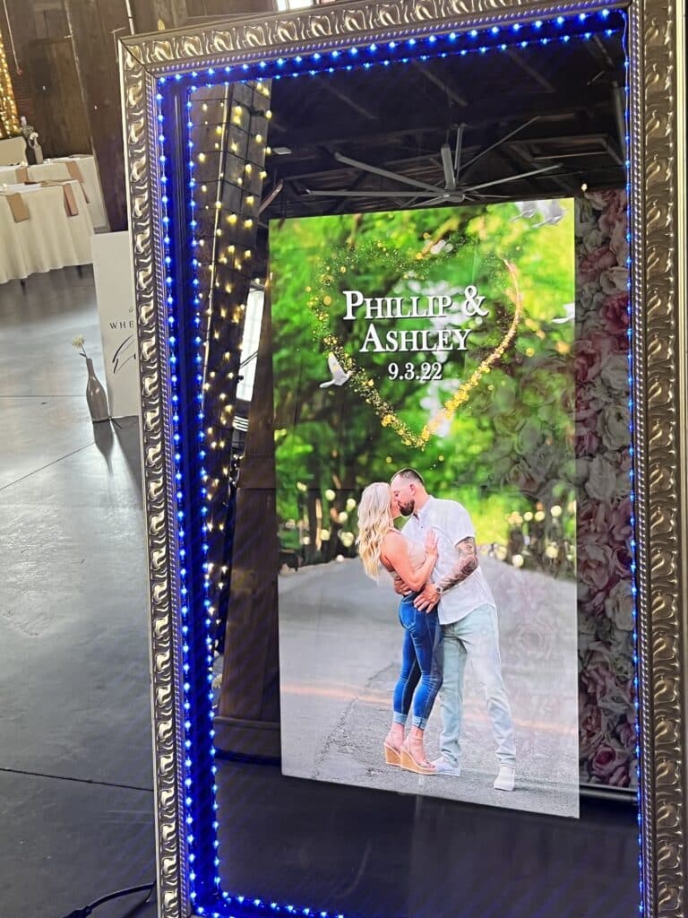 Full-length Magic Mirror touch screen photo booth with a blue LED frame showing a couple photo with a custom overlay