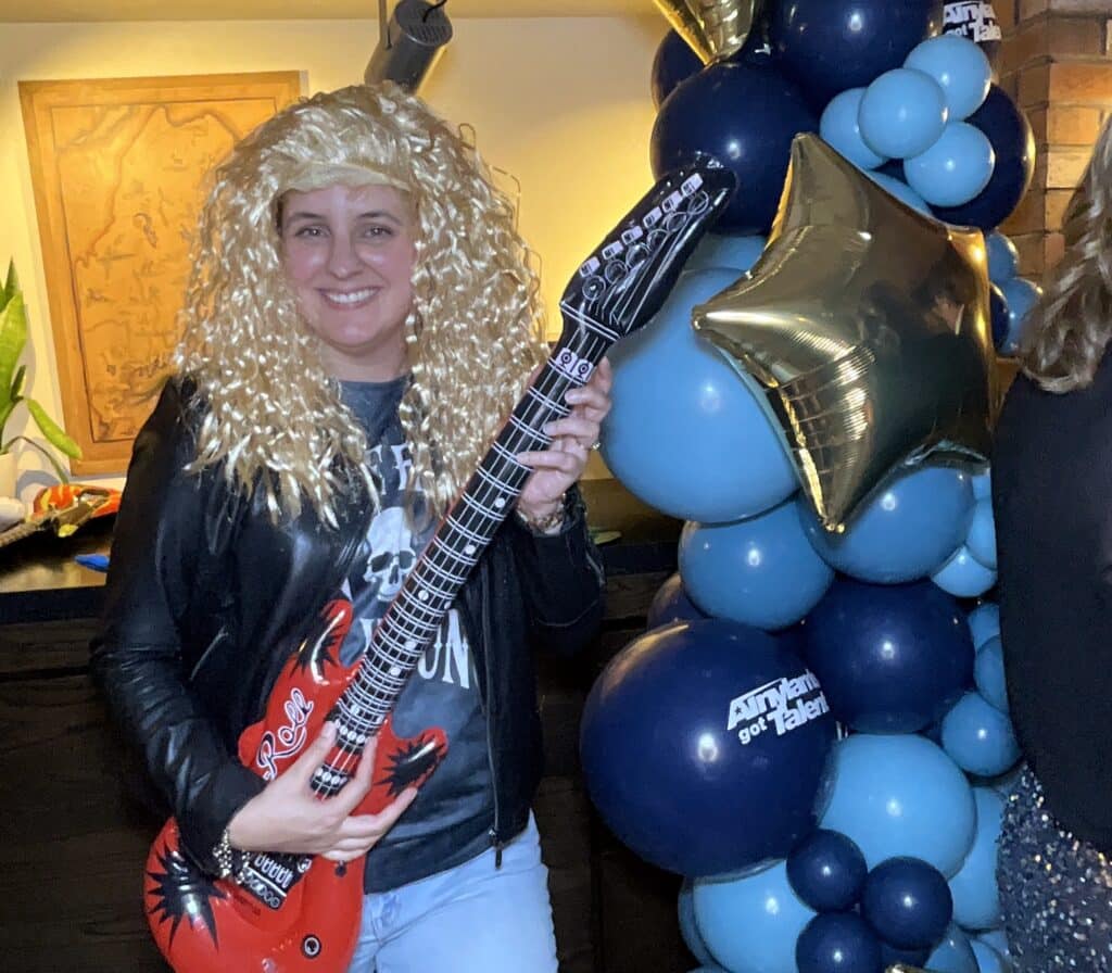 Guest in a blonde wig and leather jacket with an inflatable red guitar at a themed party at Cliff House Maine
