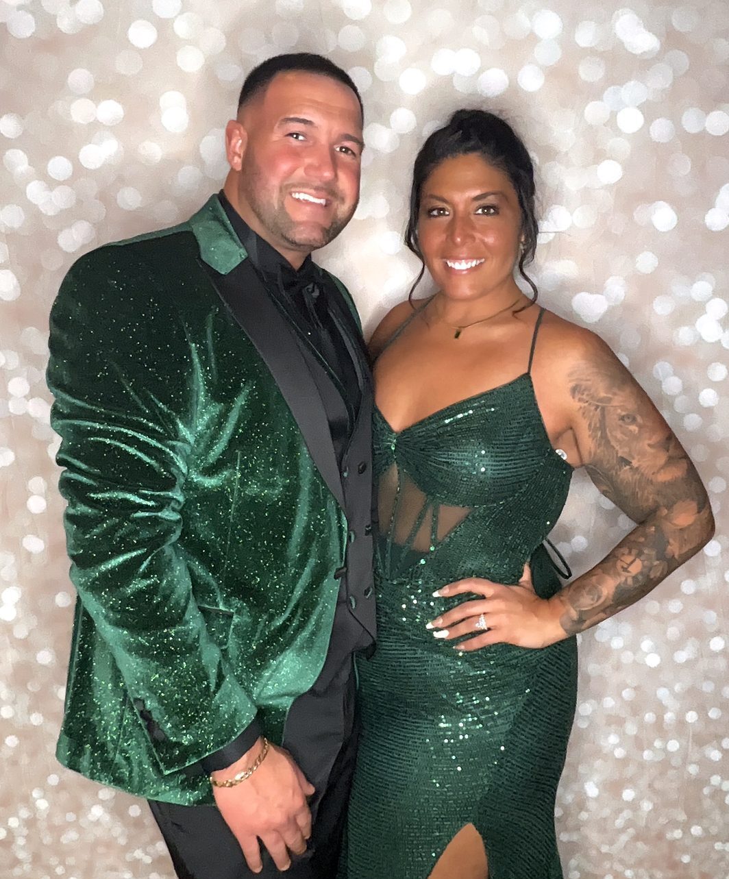 A couple in matching emerald green sparkle outfits smiling in front of a champagne glitter backdrop