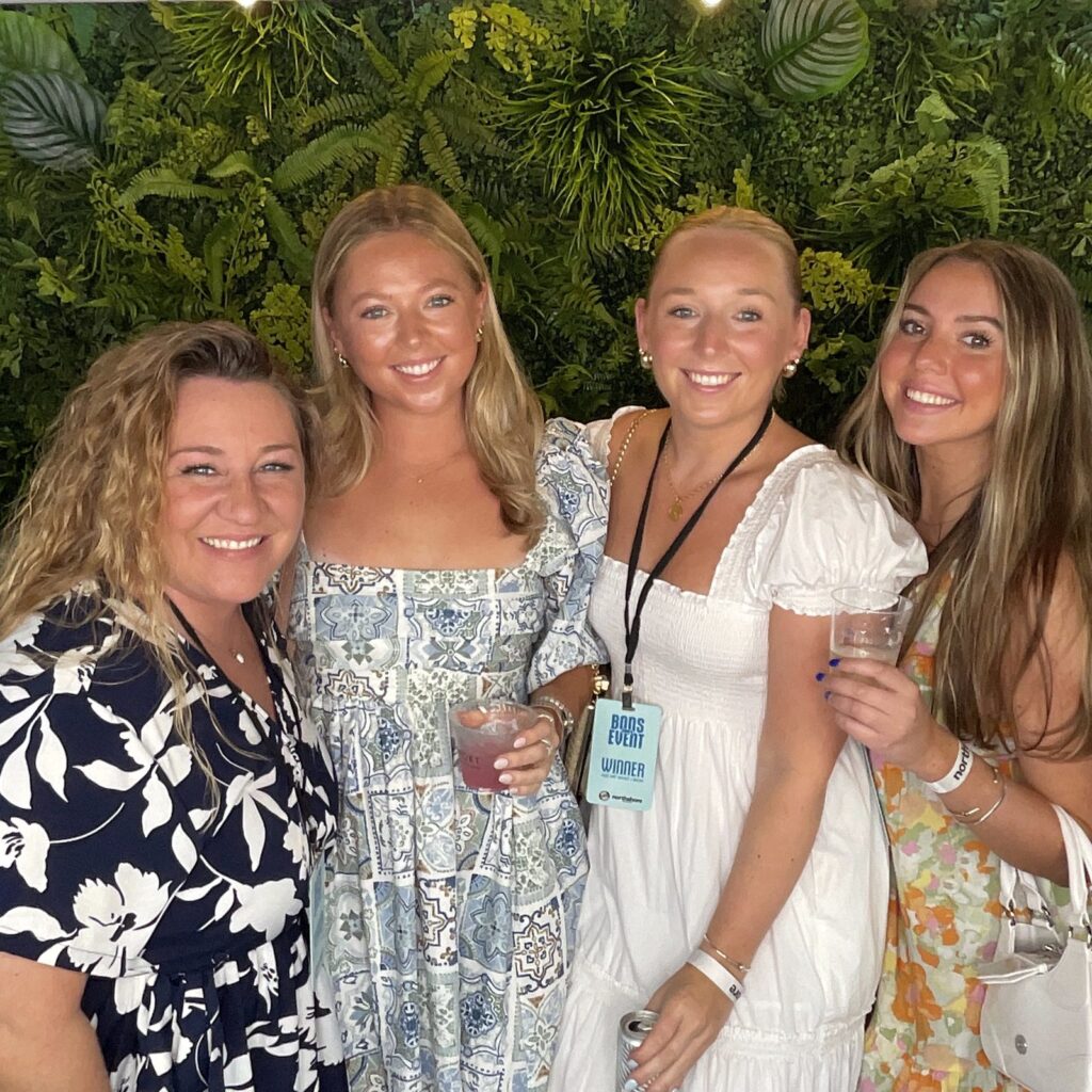 Four friends in summer dresses smiling together in front of a green foliage wall at a rooftop event