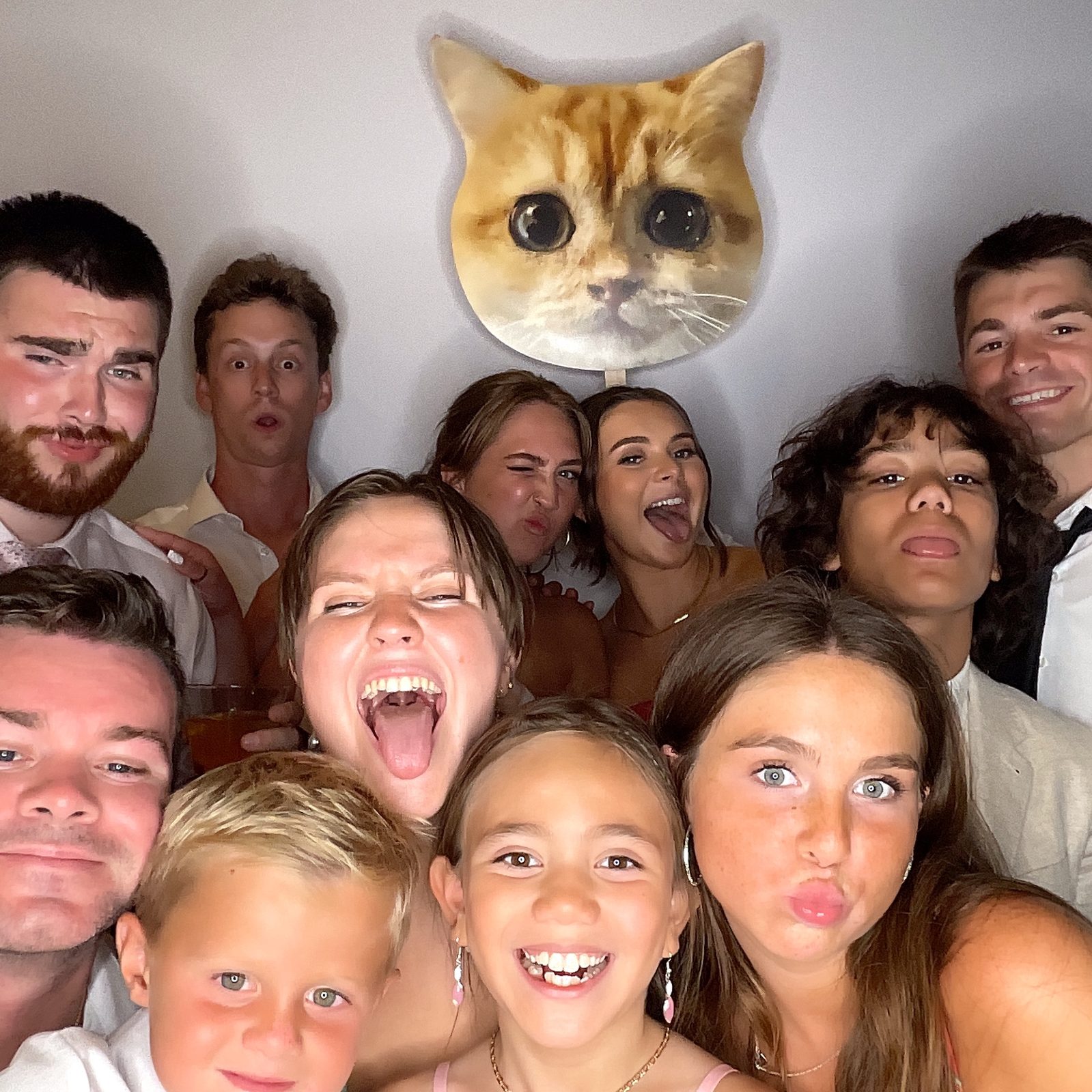A large group of wedding guests of all ages making silly faces in front of a cat head prop