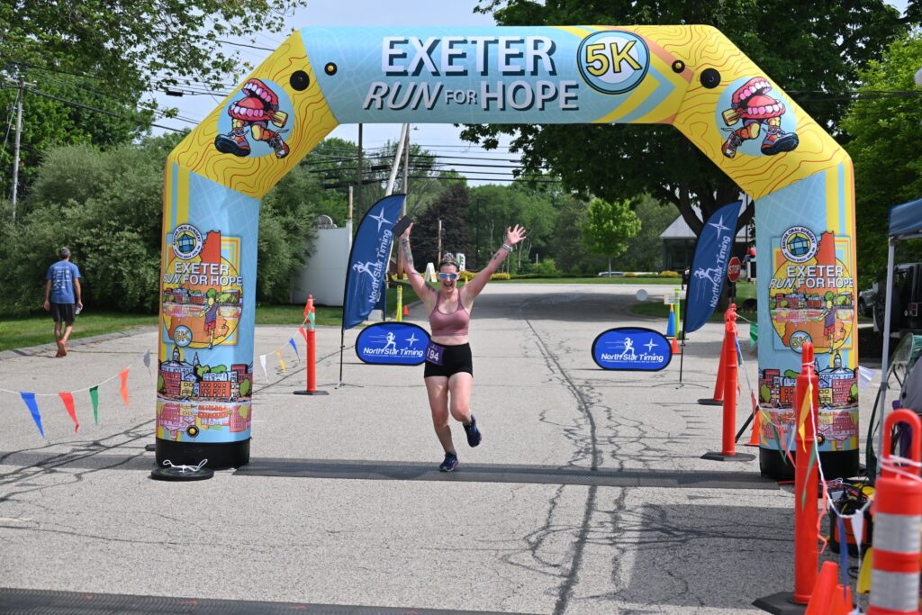Runner with arms raised celebrating under the Exeter Run for Hope finish arch in Exeter, NH