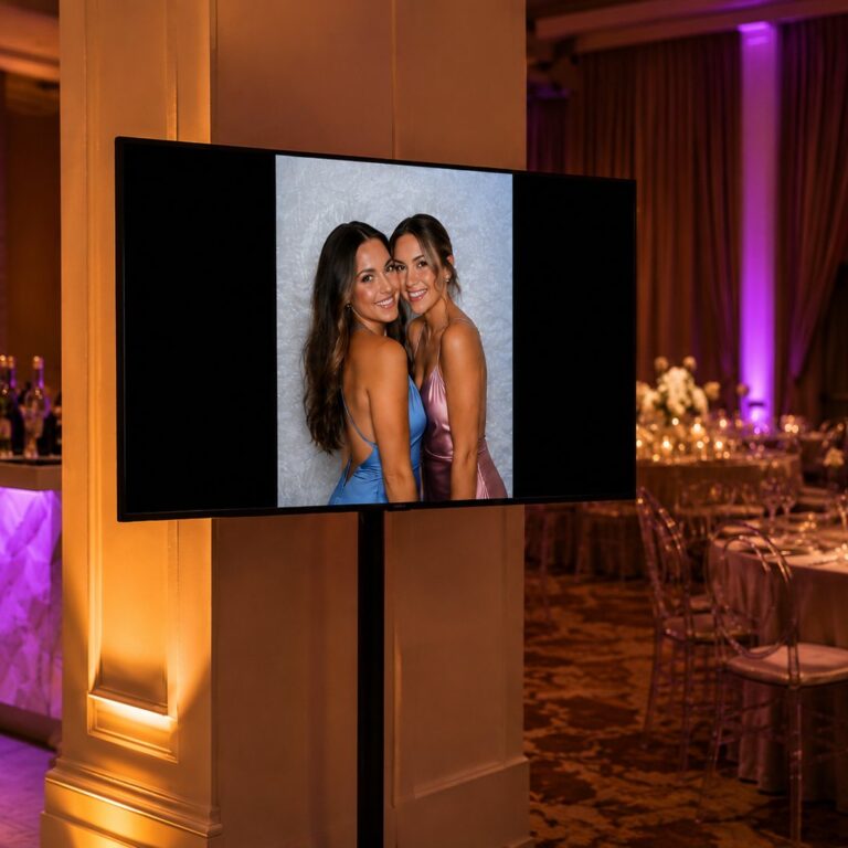 A wall-mounted TV on a stand showing a live slideshow of two friends posing in their guest photo at an evening reception