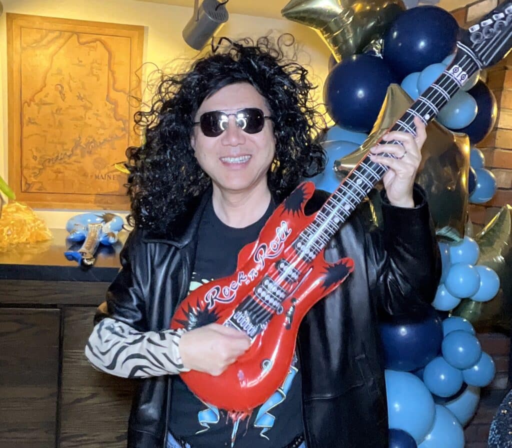 Guest in a black wig and sunglasses playing a red inflatable guitar for the roaming photo booth at a Southern Maine event