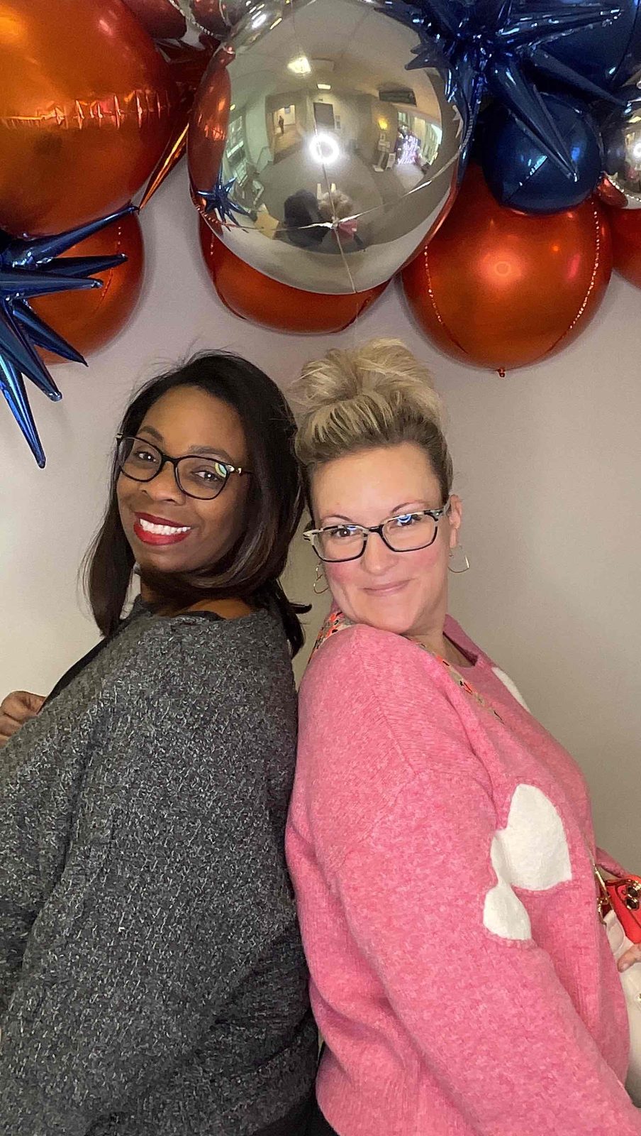 Two women standing back to back and smiling under an orange, navy and silver balloon garland