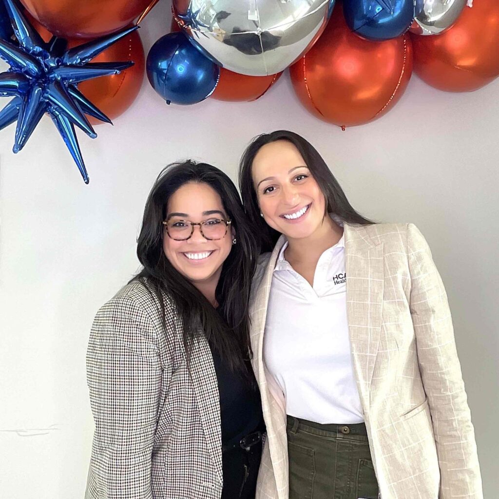 Two smiling women in blazers standing under an orange, navy and silver balloon garland