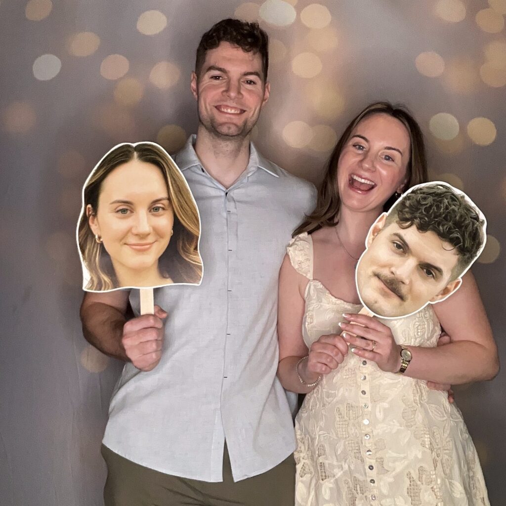 Couple smiling in front of a soft gold bokeh light backdrop, each holding a large cutout face on a stick